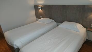 A hotel room with two single beds, a wooden floor, a wall-mounted light fixture, and a small blue accent piece on the wall.