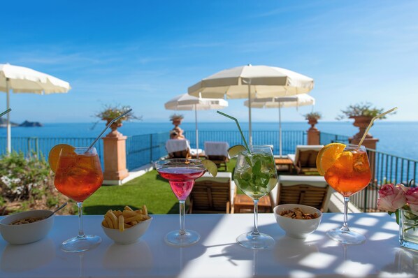 A table with four drinks and bowls of snacks, overlooking a pool and the sea.