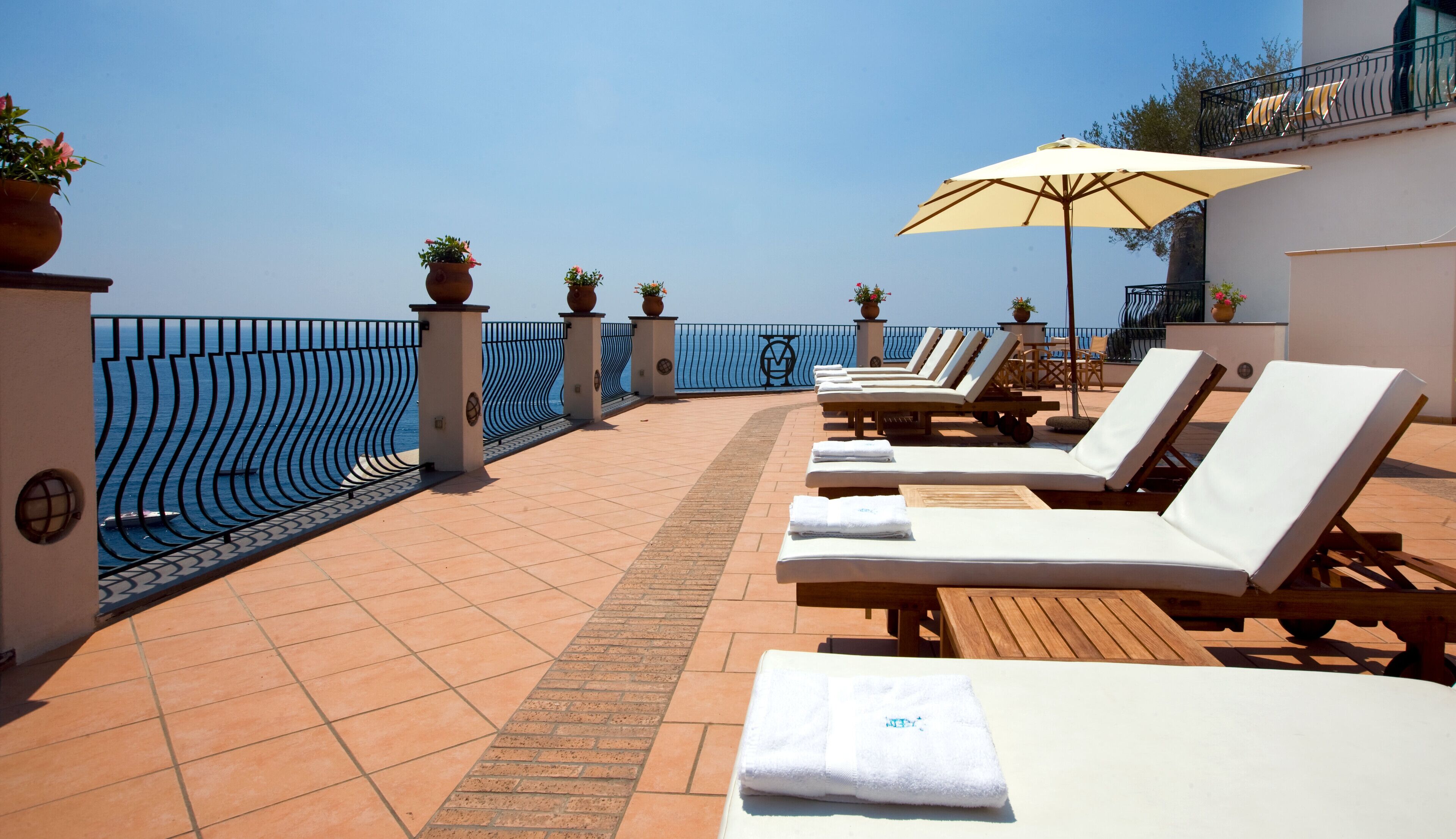 A sunlit outdoor seating area with lounge chairs and umbrellas, overlooking a clear blue sea.
