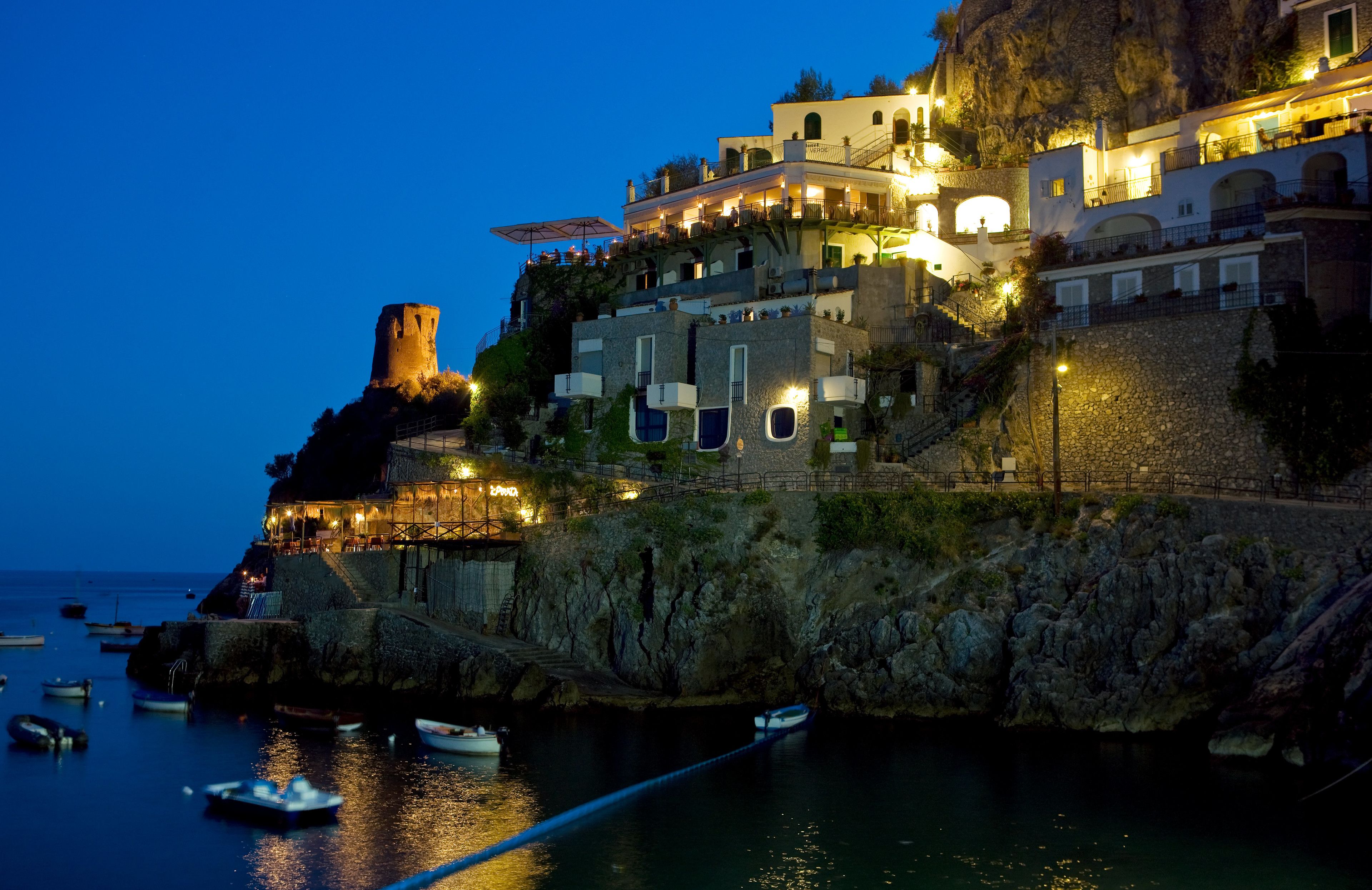 Un pueblo costero con una torre histórica, edificios escalonados y barcos en el puerto.