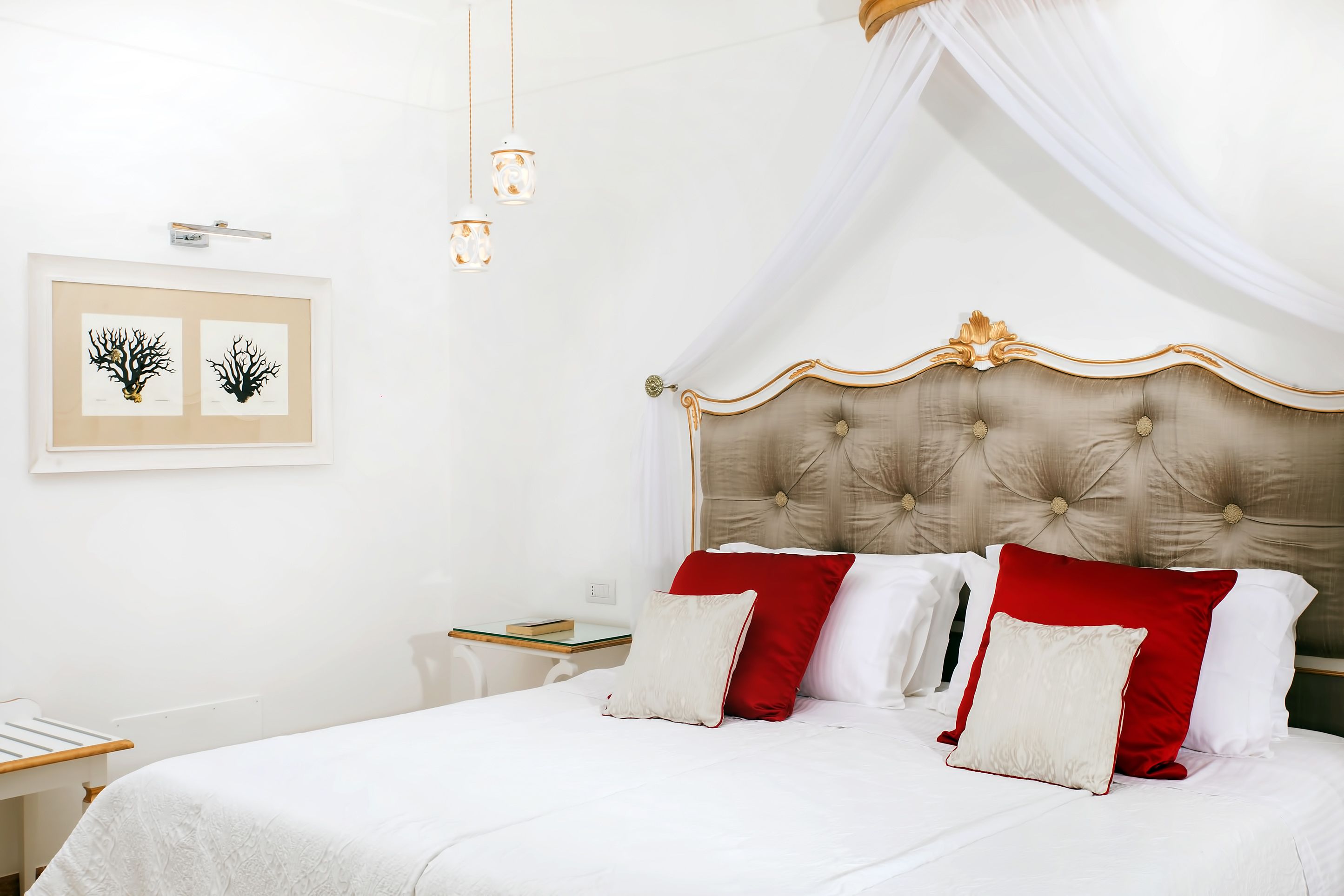 A neatly made bed with a tufted headboard, white bedding, and red and white pillows. There are framed botanical prints on the wall, a bedside table with a book, and a hanging light fixture.