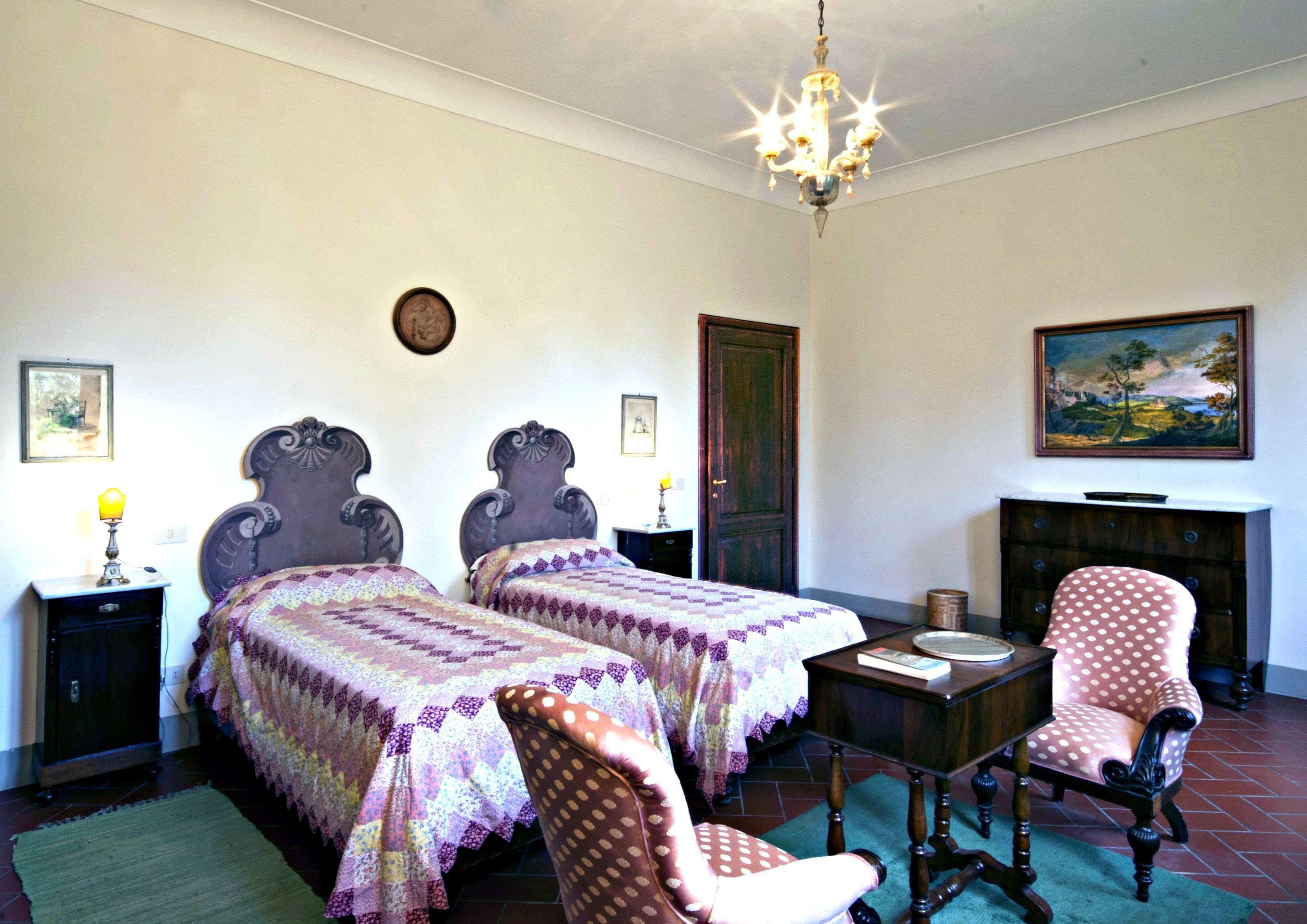 A room with two beds, a chandelier, a desk, a chair, a dresser, and a painting on the wall.
