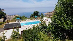Un espace piscine offrant une vue sur l’océan, entouré de verdure et bordé d’un bâtiment blanc.