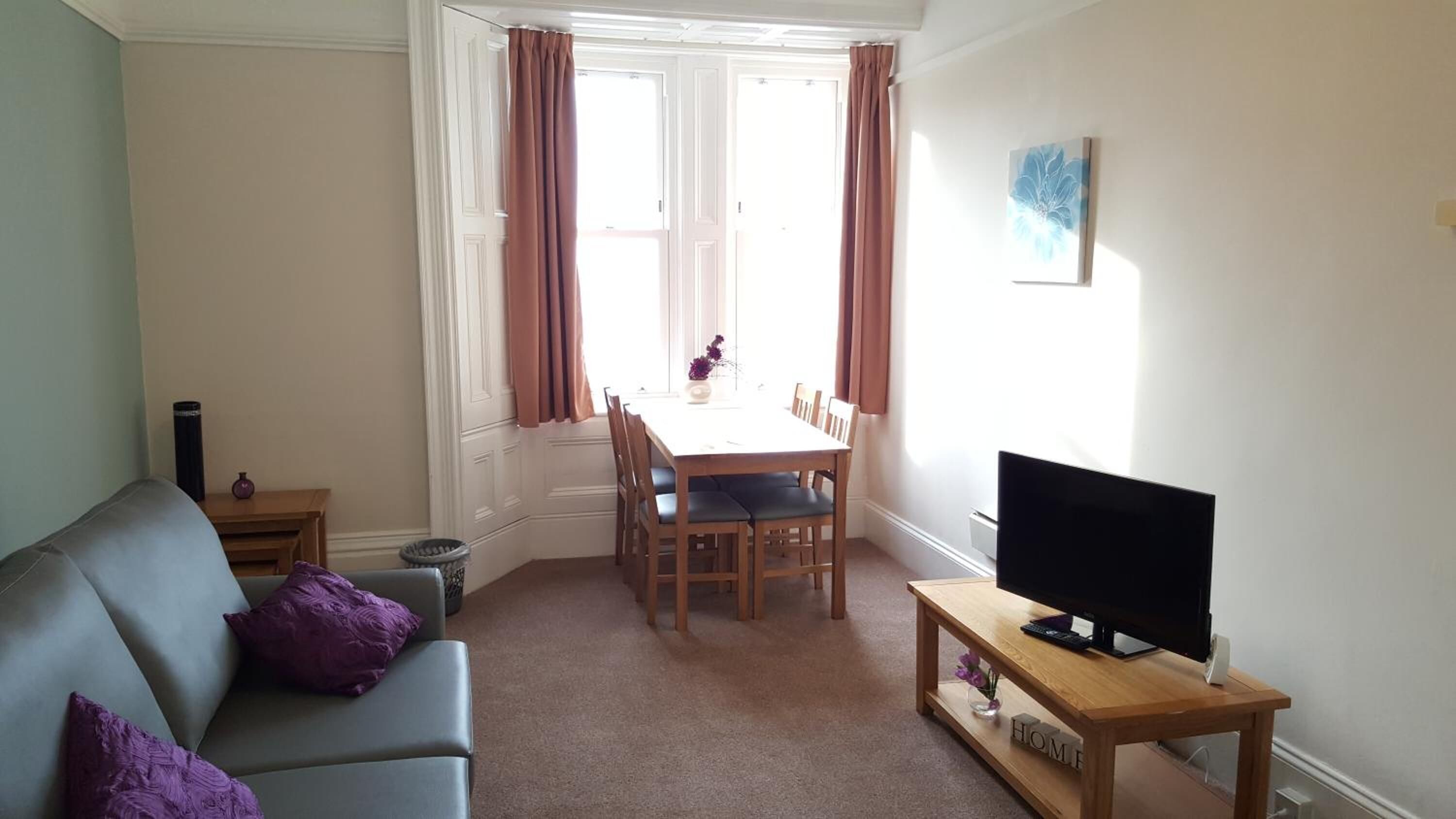 A living room with a sofa, a dining table with chairs, a television on a wooden stand, and a window with curtains.