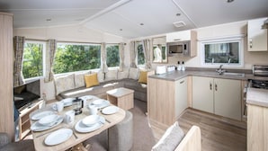 Une cuisine moderne et compacte pour camping-car, dotée d’un coin repas, d’appareils électroménagers intégrés et de grandes fenêtres offrant une vue sur la verdure.