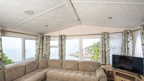 A modern living room with a sectional sofa, a flat-screen TV, and large windows offering a view of the sea and cliffs. - Sandaway Beach Holiday Park (Ilfracombe)