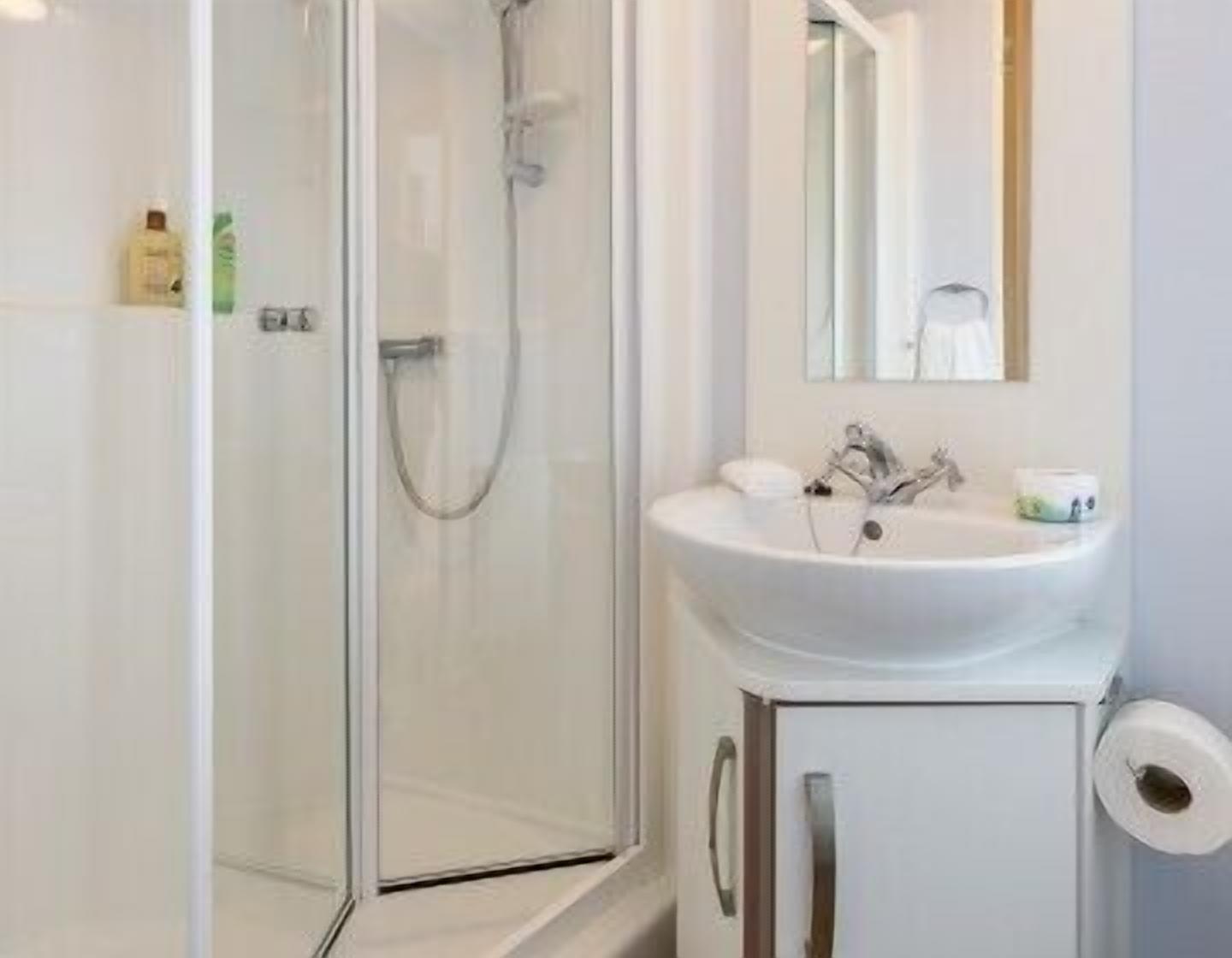 Uma casa de banho com cabine de chuveiro em vidro, uma pia branca com espelho em cima e uma sanita com suporte para toalhas de papel.