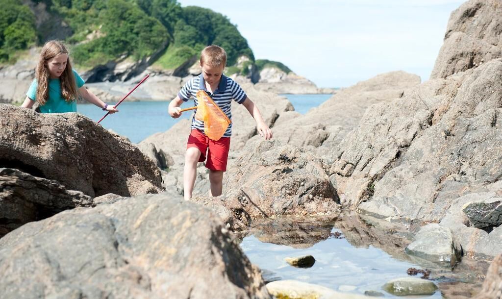 Two children playing near rocky terrain with a body of water and greenery in the background.