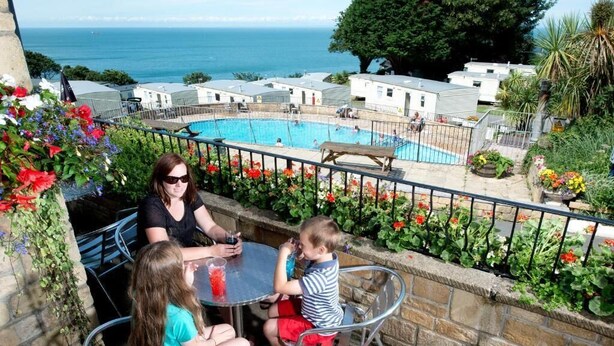A poolside area with a view of the sea, a dining table, and a child playing. - Sandaway Beach Holiday Park (Ilfracombe)