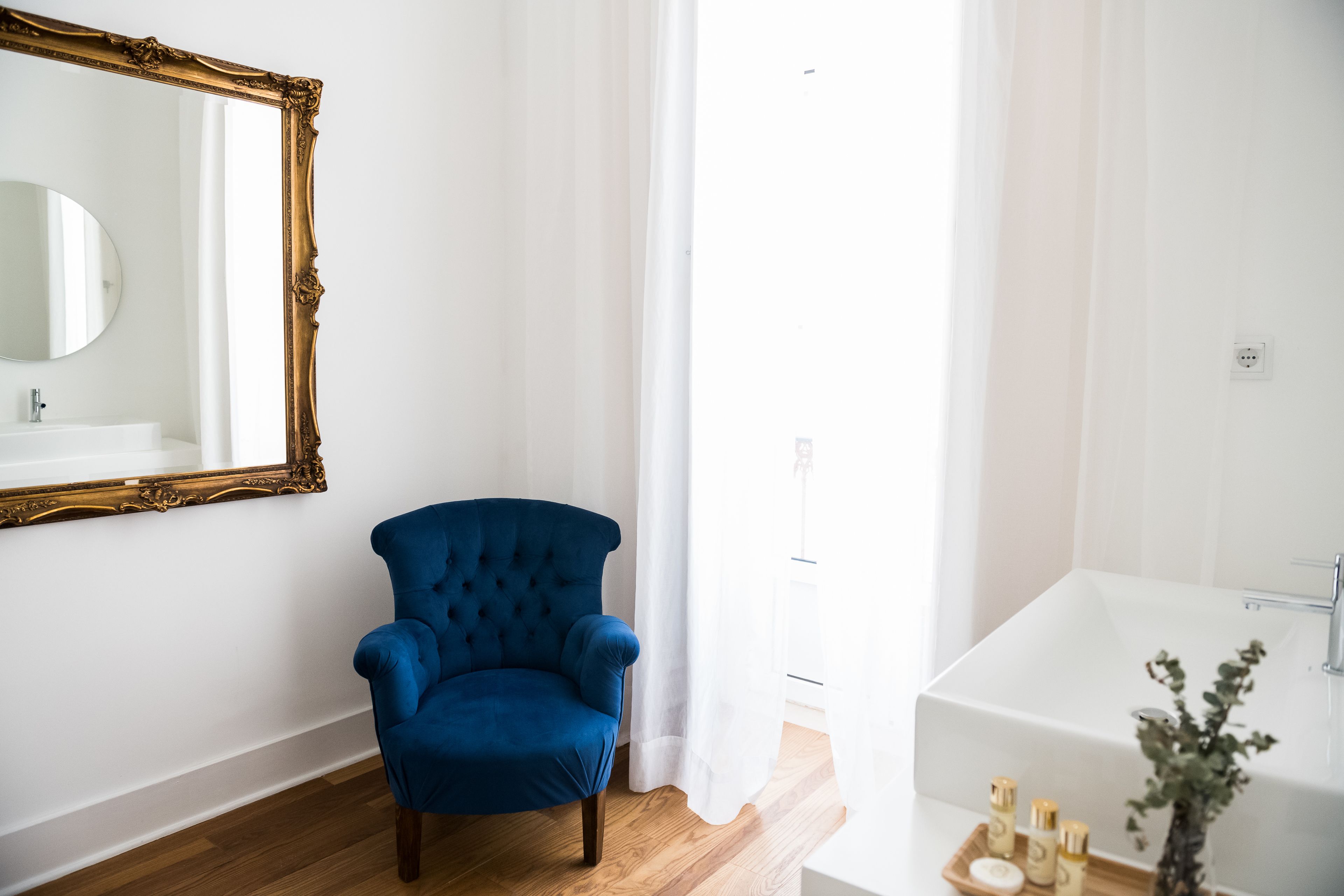 A bathroom with a large mirror, a blue armchair, a white bathtub, and a wooden floor.