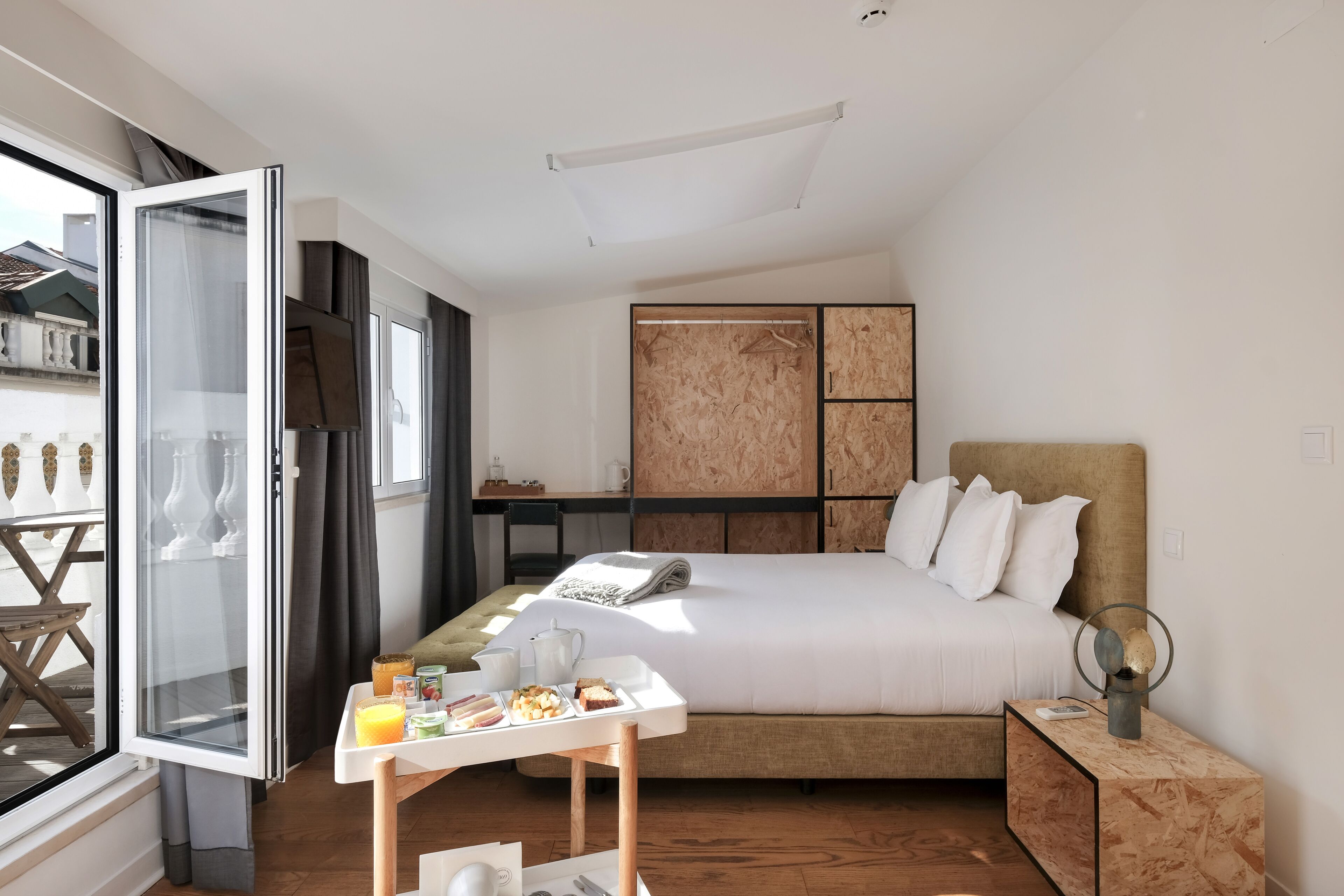 Una habitación de hotel moderna con una cama grande, un escritorio con provisiones para el desayuno, un armario y vistas a través de una puerta abierta.