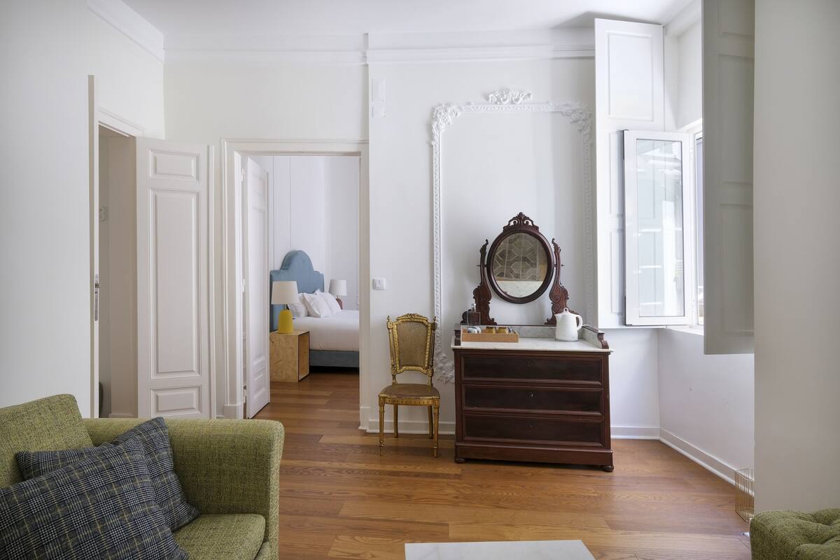 Une pièce avec un canapé vert, une commode en bois, un miroir et un lit à l’arrière-plan.