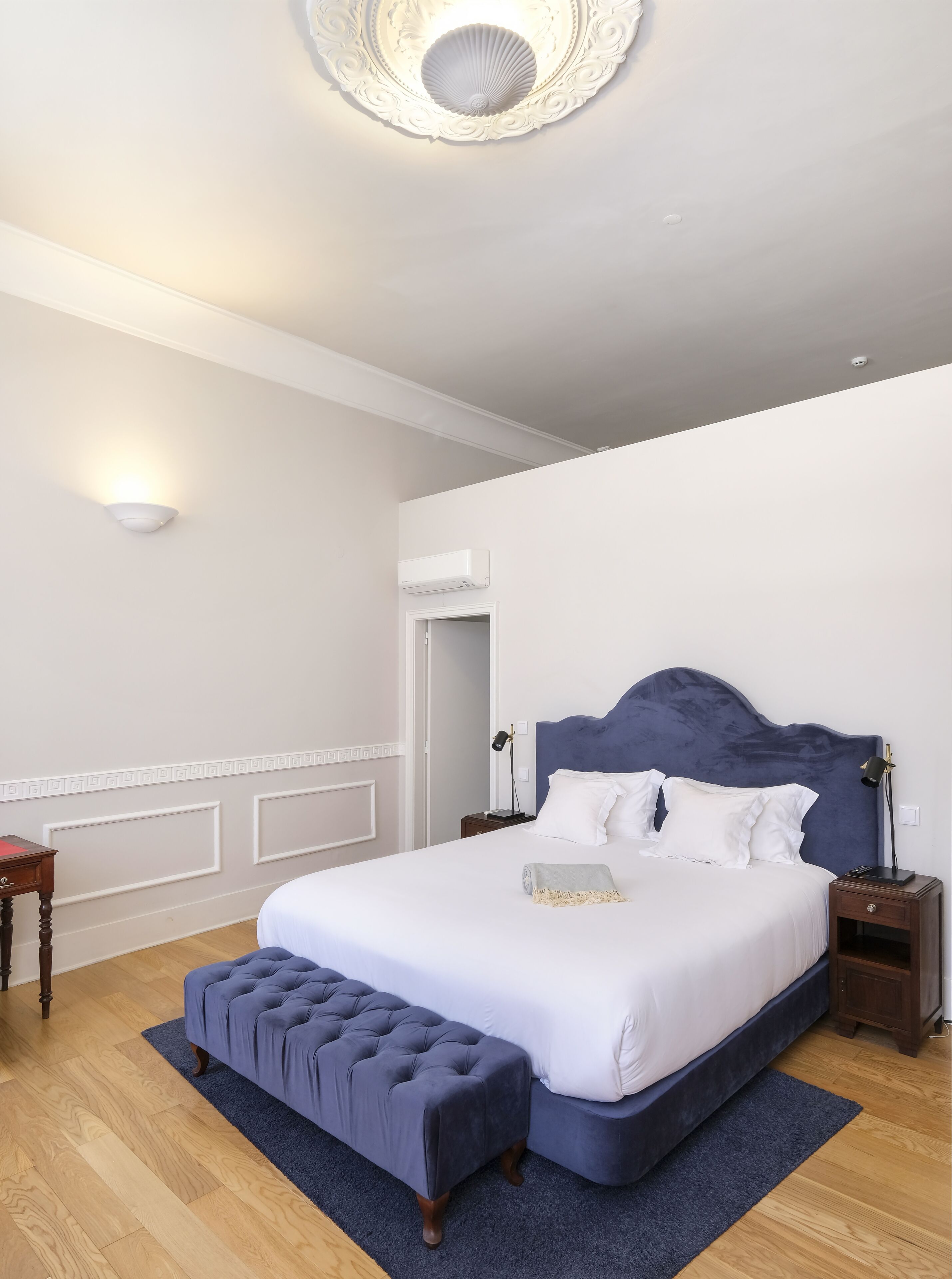 Une chambre à coucher avec un grand lit, une table de chevet, une commode, une lampe fixée au mur et un ventilateur de plafond.