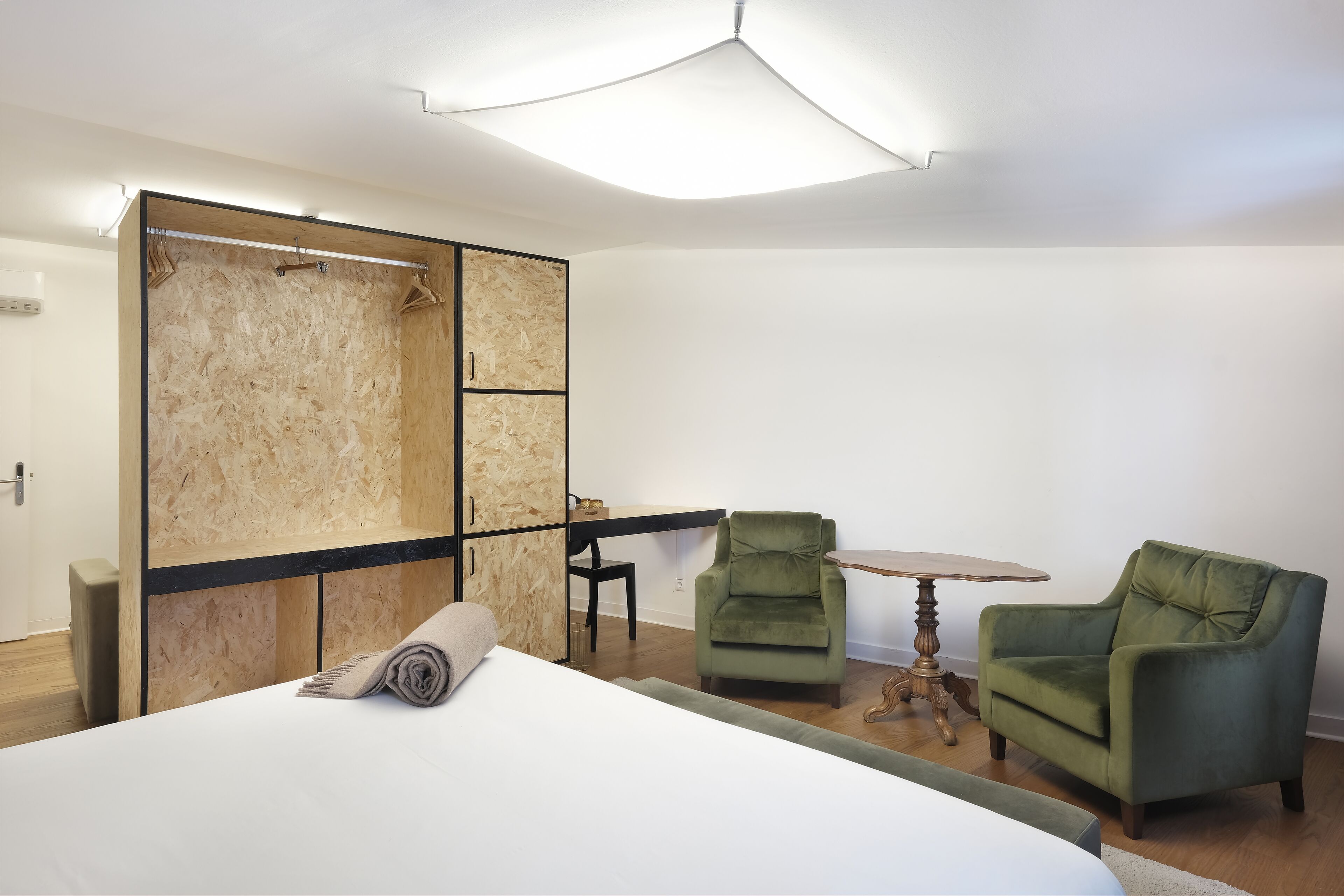 A hotel room with a bed, a wooden wardrobe, two green armchairs, a small round table, and a ceiling light fixture.