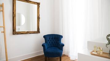 Ein Zimmer mit einem blauen Sessel, Holzboden, weißer Wand und einem großen, verzierten Spiegel.