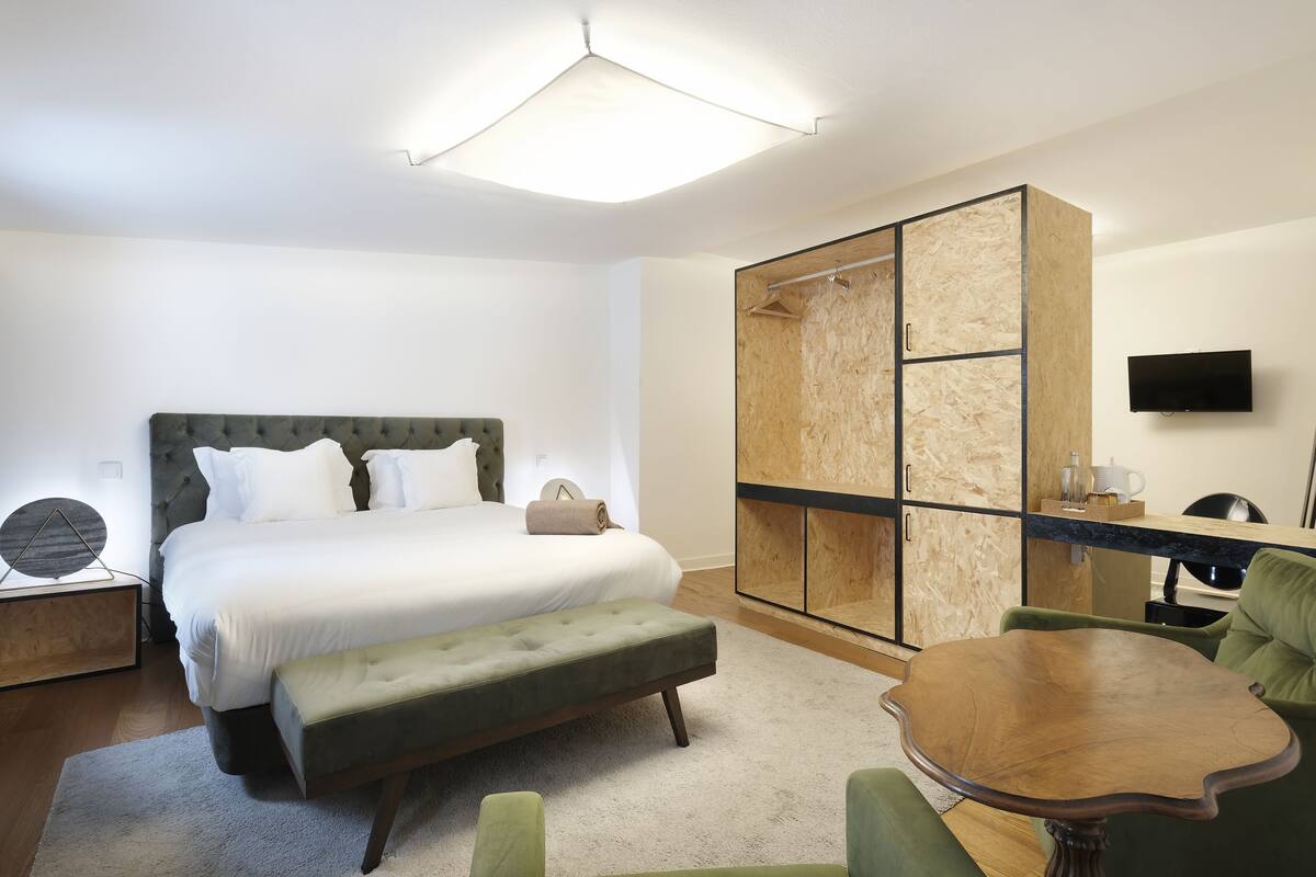 Une chambre d’hôtel moderne avec un grand lit, une armoire en bois, une petite table et un fauteuil vert.