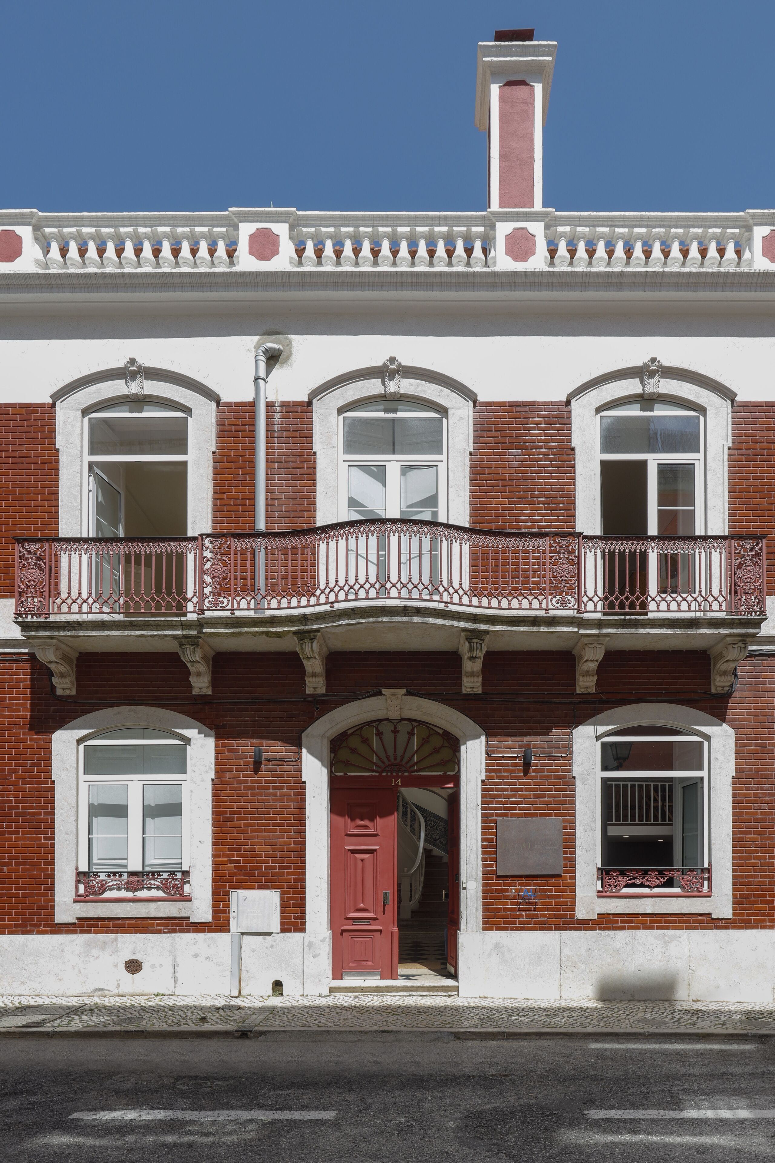 Un edificio de dos pisos con fachada de ladrillo rojo, detalles blancos y un balcón con barandilla de metal decorativo.