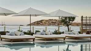 Un área junto a la piscina con sillones y sombrillas, vista al mar y una isla rocosa al fondo.