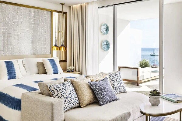 A modern hotel room with a large bed, a sofa, a small table, and a balcony with a view of the sea.