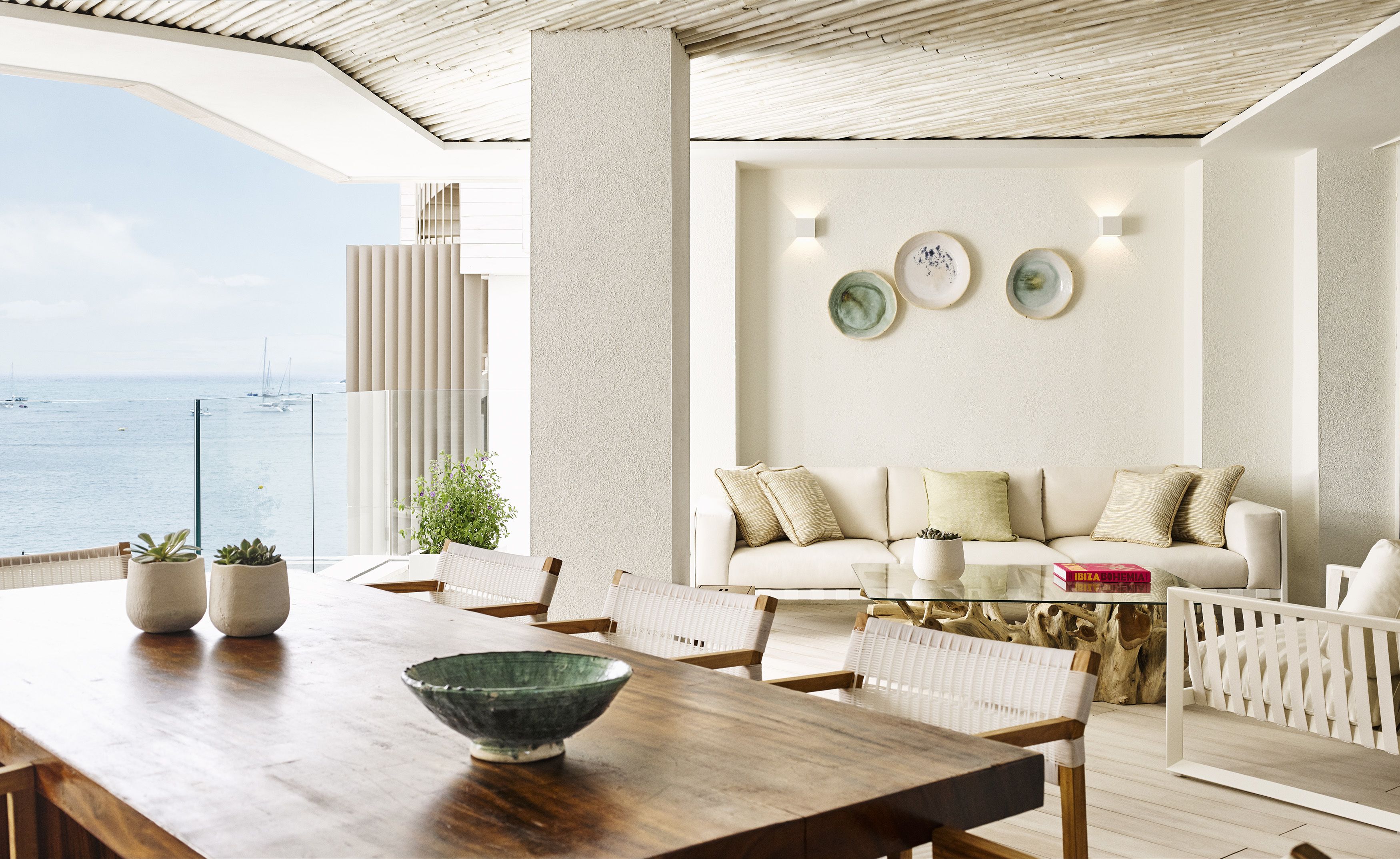 A modern living room with a view of the sea, a wooden dining table, and a sofa with cushions.