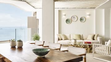 A modern living room with a view of the sea, a wooden dining table, and a sofa with cushions.