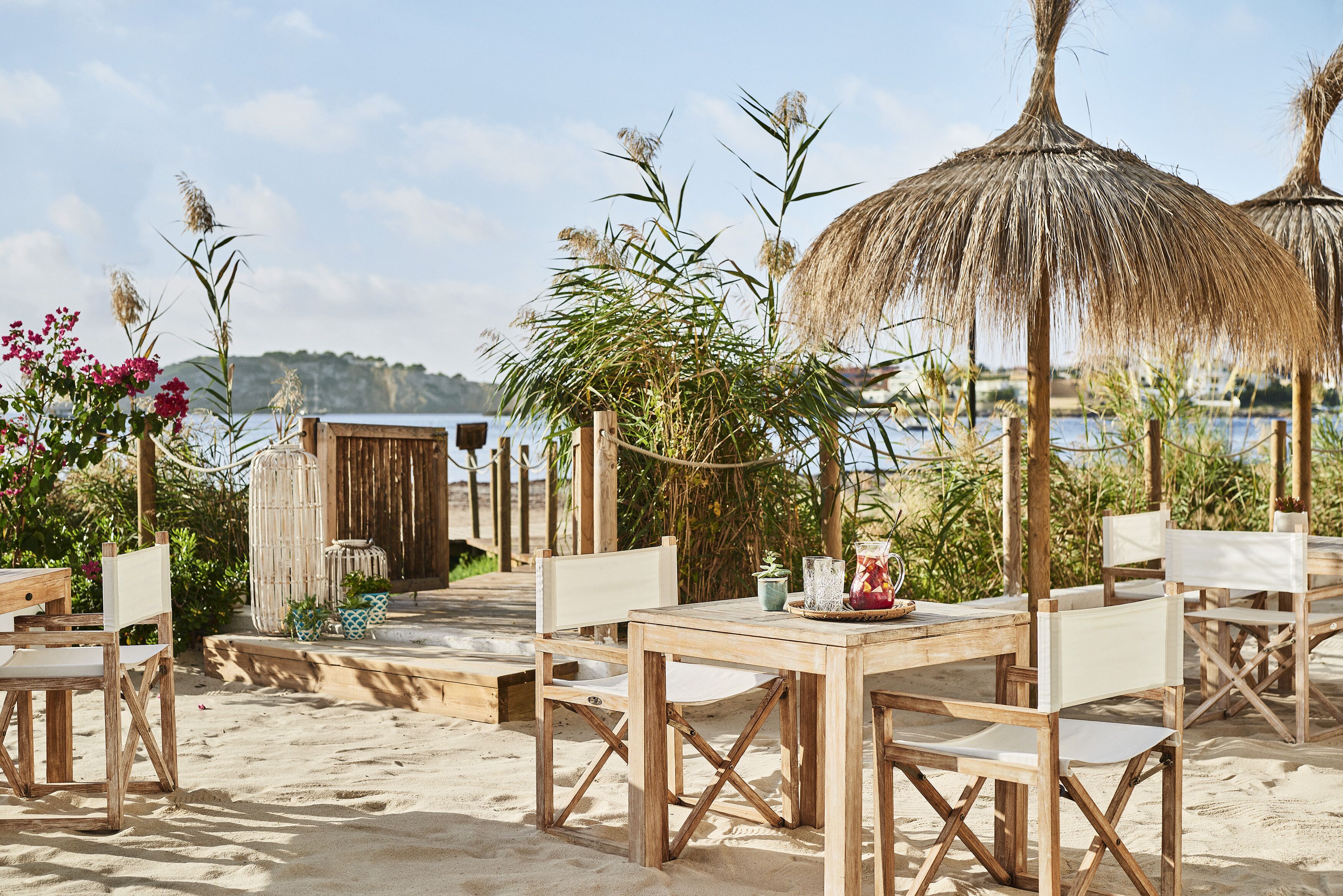 An outdoor dining area with wooden tables and chairs, surrounded by plants and a thatched umbrella.