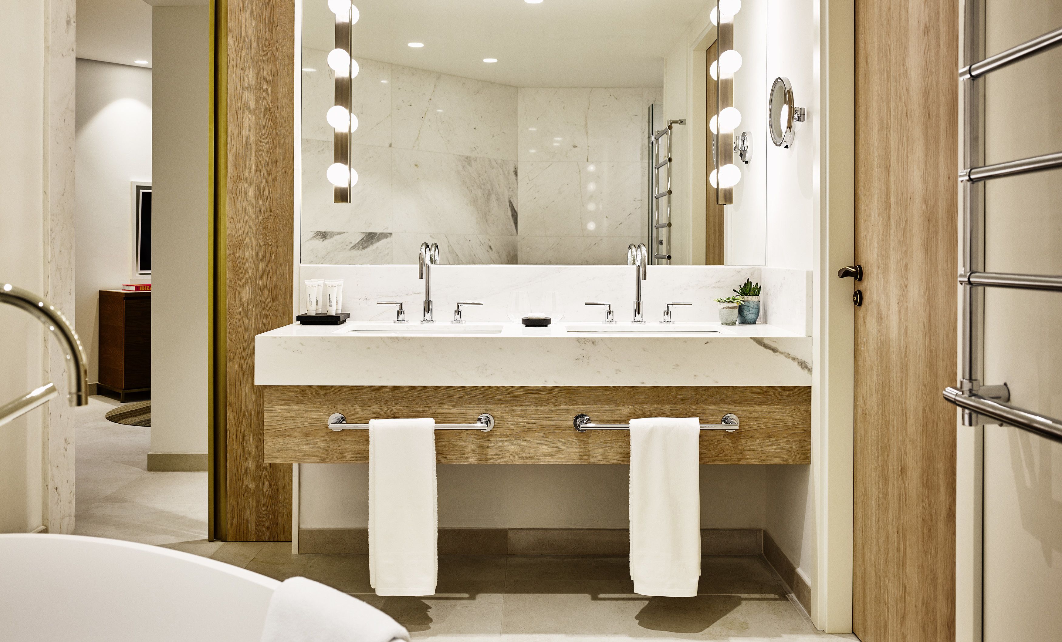 A modern bathroom with a double sink, vanity, and a large mirror with lights. There are towels hanging on the side and a small plant on the vanity.