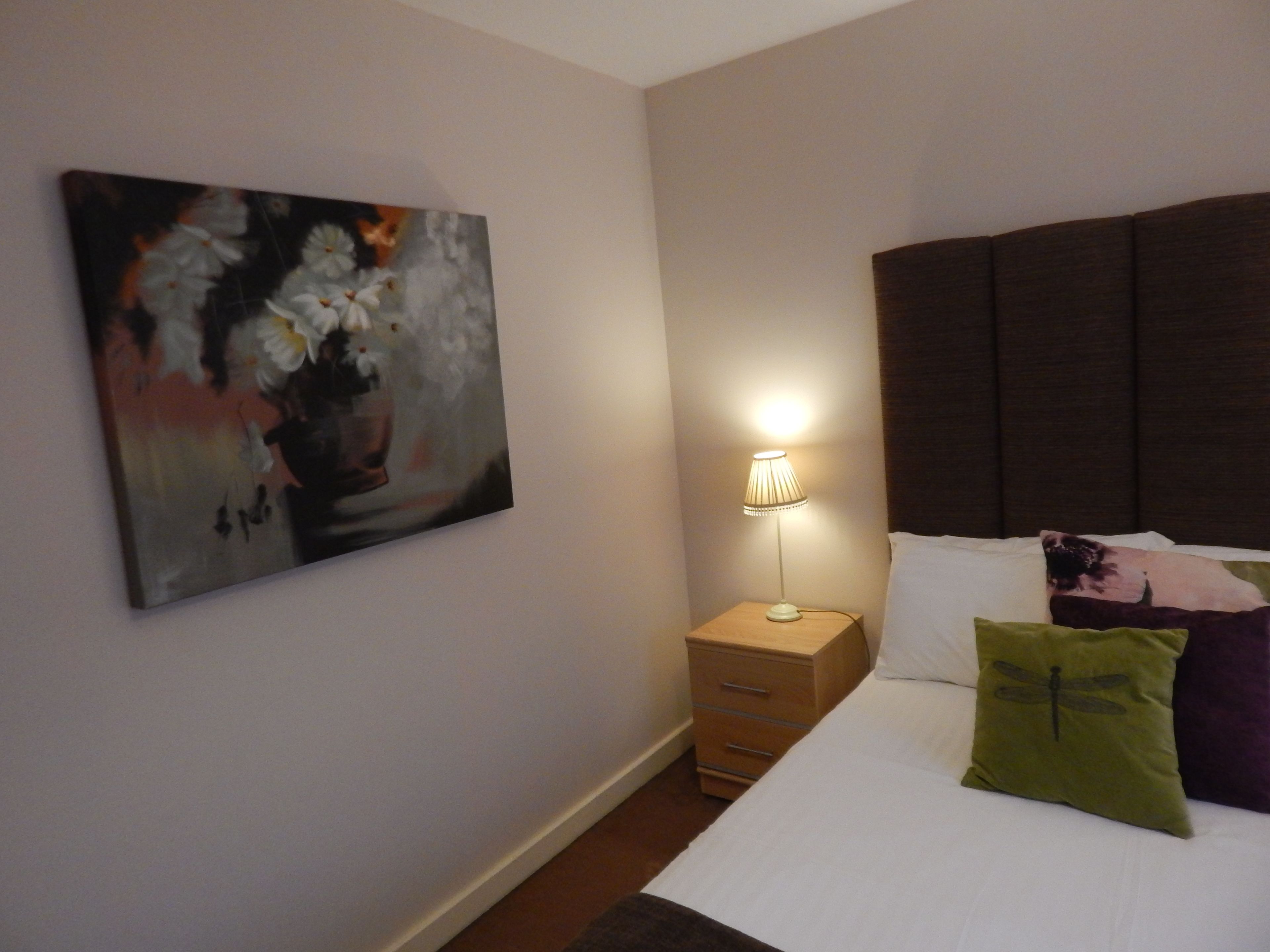 Une chambre à coucher avec un lit, une table de chevet, une lampe, un oreiller orné d’un motif de libellule et un tableau accroché au mur.