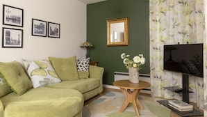 A green sofa with patterned cushions, a round wooden table with a vase of white flowers, and a black TV stand with a flat-screen TV. - LETS Quiet Cul-de-Sac Apartment (Cambridge)