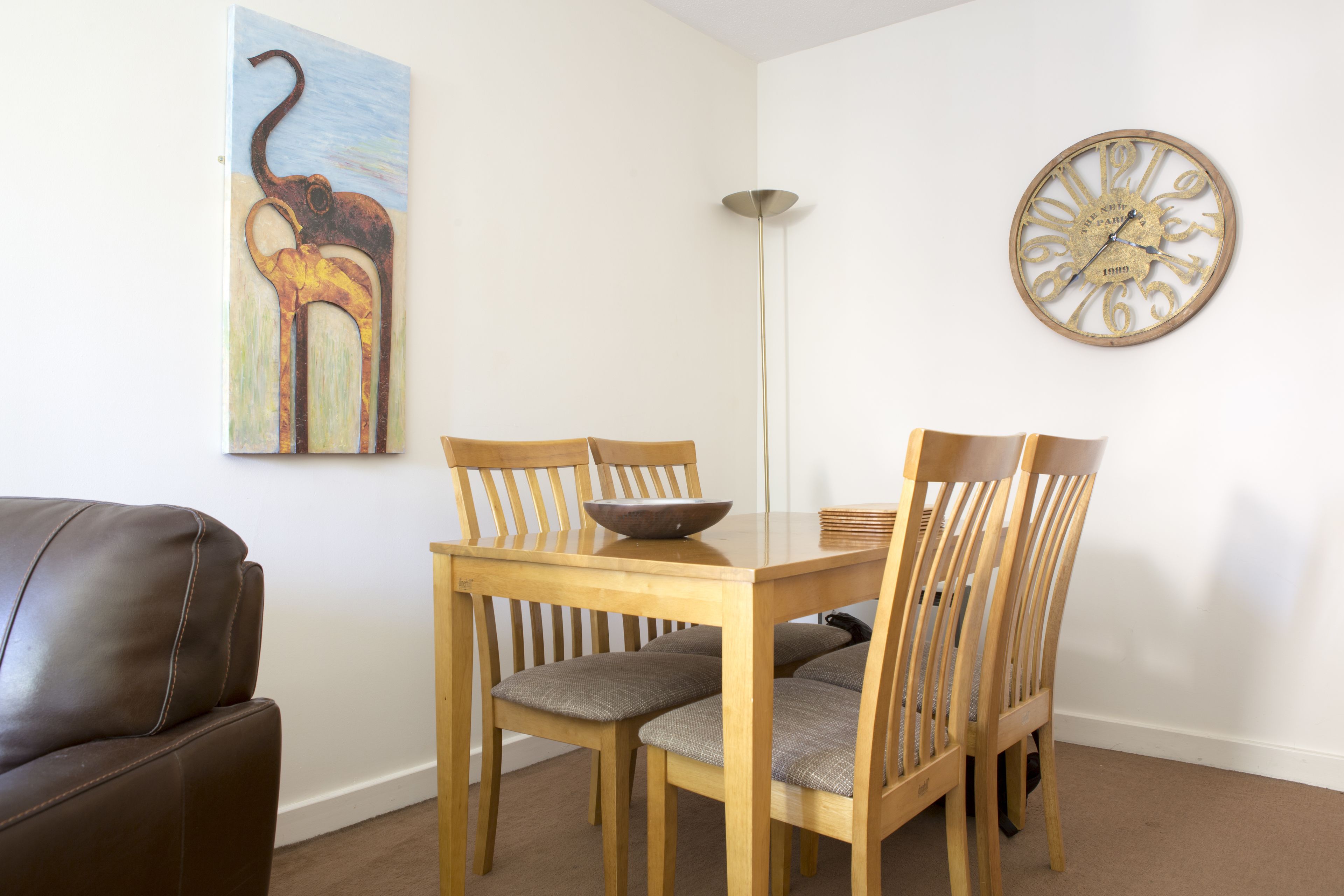 Uma sala de jantar com uma mesa de madeira e quatro cadeiras, uma tigela sobre a mesa, um relógio de parede e um quadro de um elefante.
