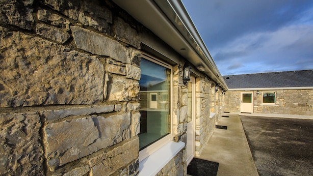A stone building with a window and a door, a concrete pathway, and a paved area. - The Red Cottage & Stables (Sligo)