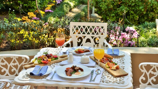 Een eettafel buiten, gedekt met ontbijtartikelen, met een weelderige tuin op de achtergrond.
