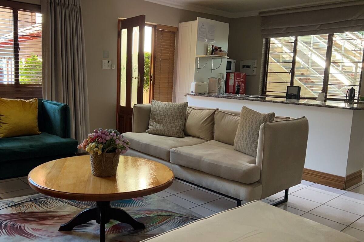 Sala de estar con un sofá verde, un sofá beige y una mesa de centro redonda con un jarrón de flores. Se observa una zona de cocina con un refrigerador y un microondas.