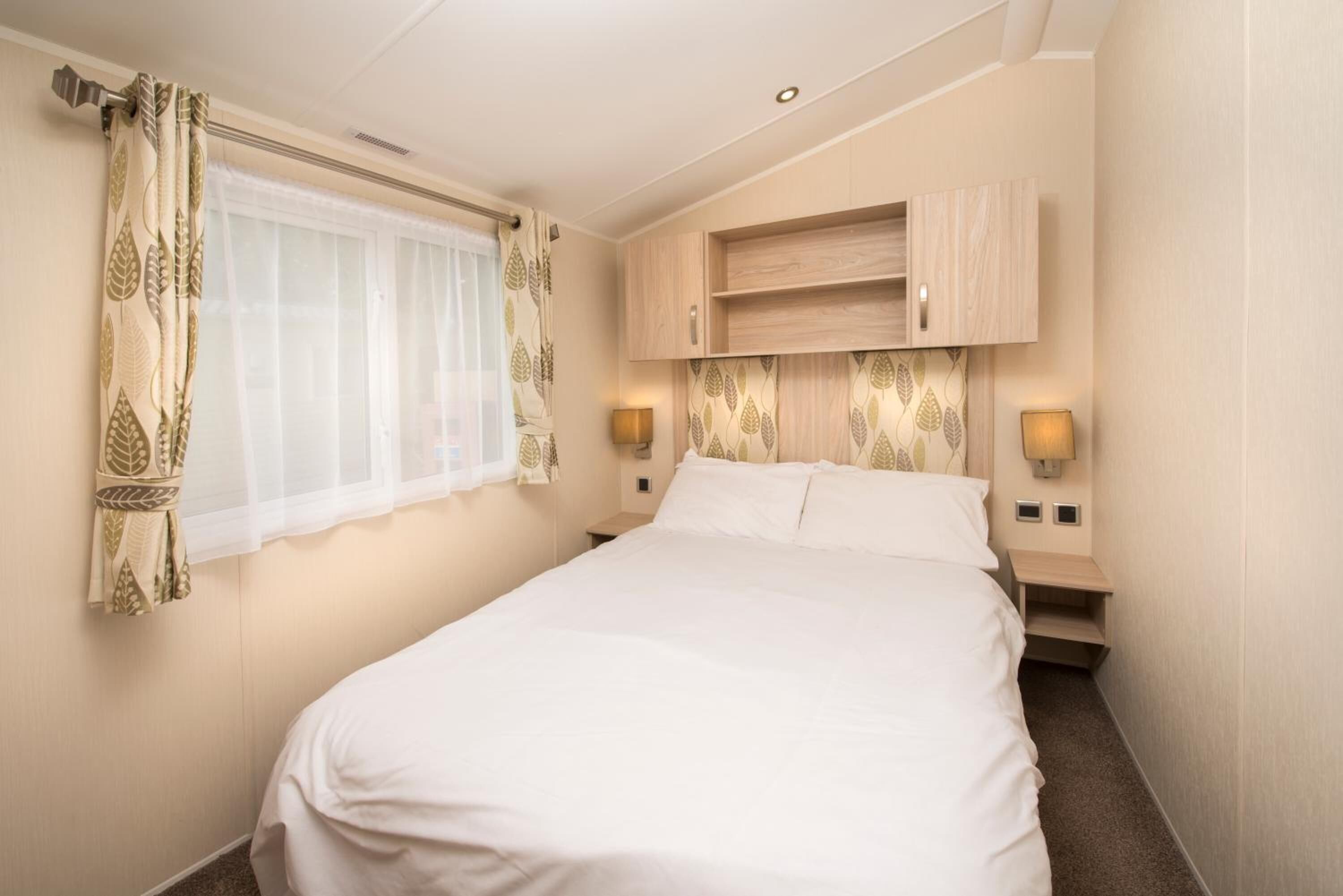 A hotel room with a bed, a window with sheer curtains, a bedside table with a lamp, and built-in wooden cabinets above the bed.