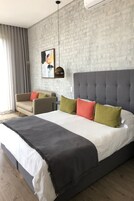 A modern hotel room with a large bed, a grey upholstered headboard, and a grey brick-patterned wall.