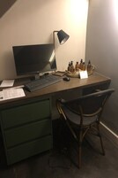Et skrivebord med en computer, tastatur, mus, lampe og en bakke med flasker. Der ligger papirer på skrivebordet, og en stol står foran det.