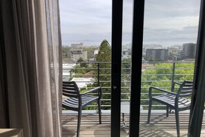 A balcony with two metal chairs and a table, overlooking a cityscape with buildings and greenery.