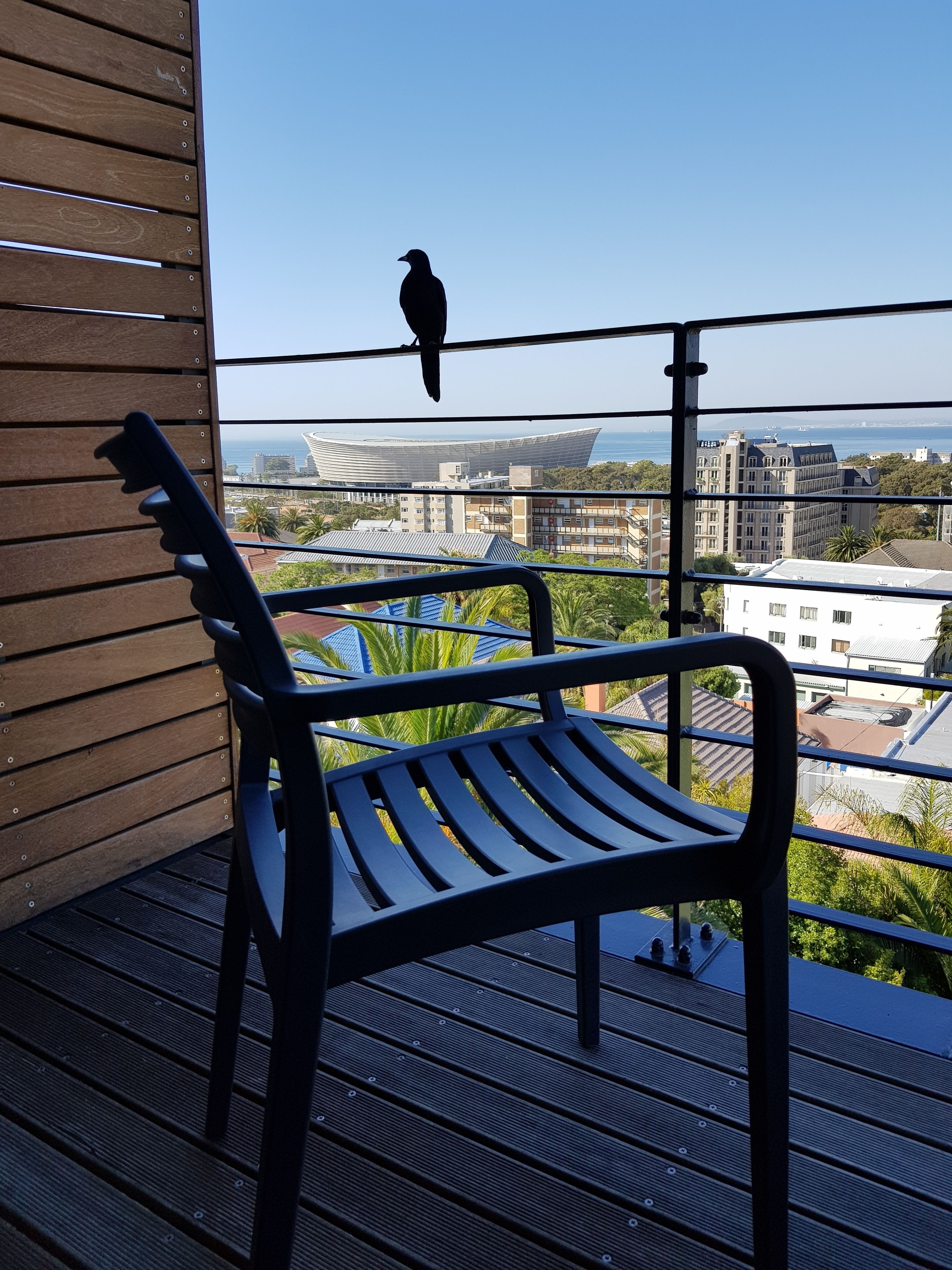 Ein Balkon mit Holzstuhl und Metallgeländer mit Blick auf eine Stadtlandschaft, einschließlich Gebäude und einem Gewässer.