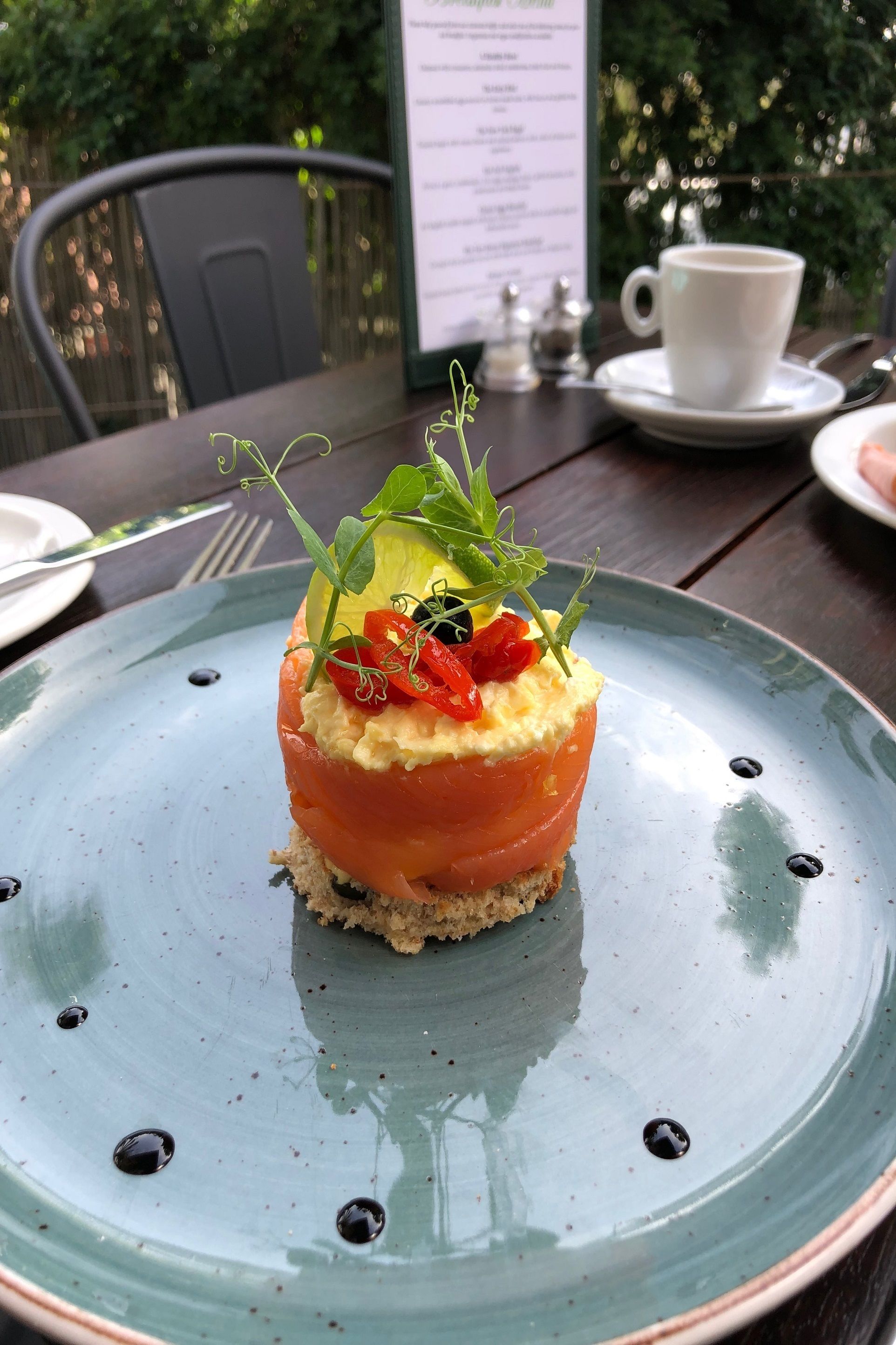Une assiette avec une entrée de tartare de saumon, une tasse de café et un menu sur une table à l’extérieur.