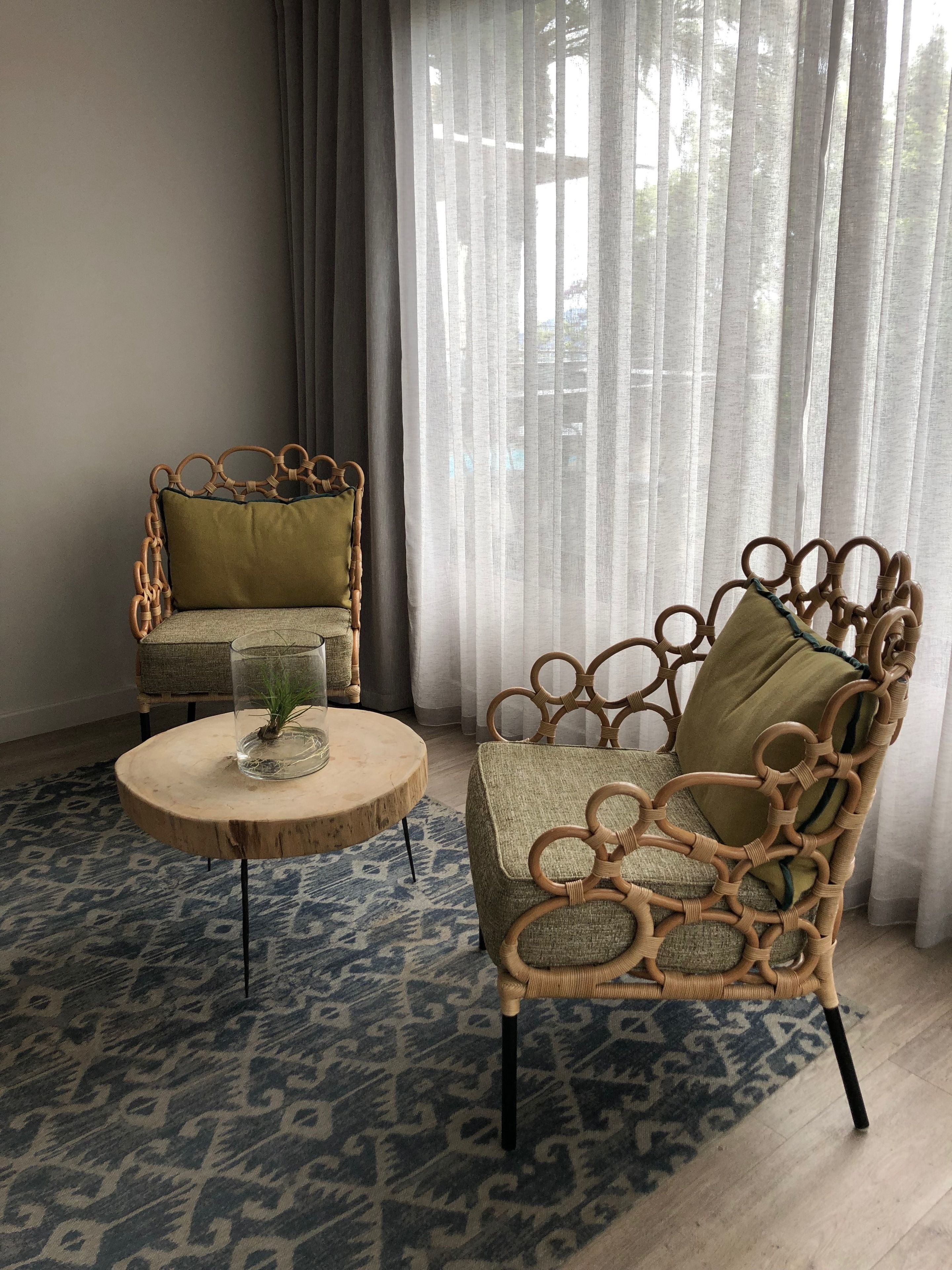 Une pièce comprenant deux fauteuils en osier, l’un avec un coussin vert et l’autre avec un coussin beige, une petite table ronde en bois sur laquelle se trouvent un vase en verre et une plante, ainsi qu’un tapis bleu à motifs.