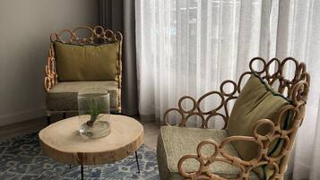 Une pièce comprenant deux fauteuils en osier, l’un avec un coussin vert et l’autre avec un coussin beige, une petite table ronde en bois sur laquelle se trouvent un vase en verre et une plante, ainsi qu’un tapis bleu à motifs.