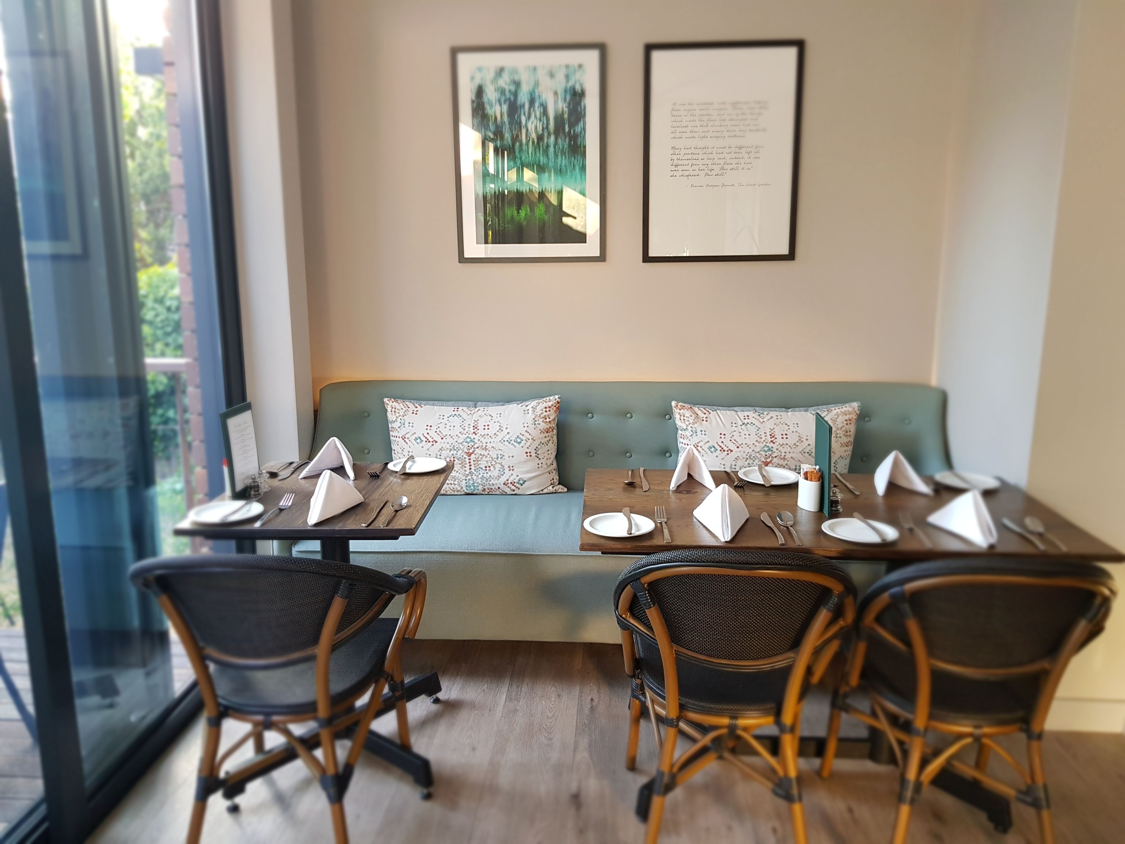 A dining area with a green sofa, two dining tables set with plates, cutlery, and napkins. There are wicker chairs, a window with a view of greenery, and two framed pictures on the wall.
