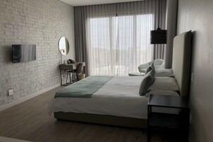 A modern hotel room with a large bed, a desk with a chair, a television, and a large window with curtains.