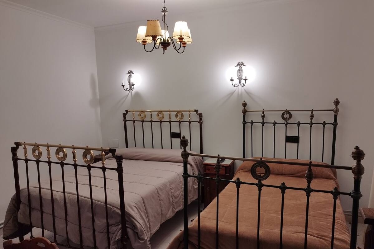 Une chambre avec deux lits, chacun doté d’un cadre en métal et d’une tête de lit. On y trouve également une chaise en bois et une petite table en bois.
