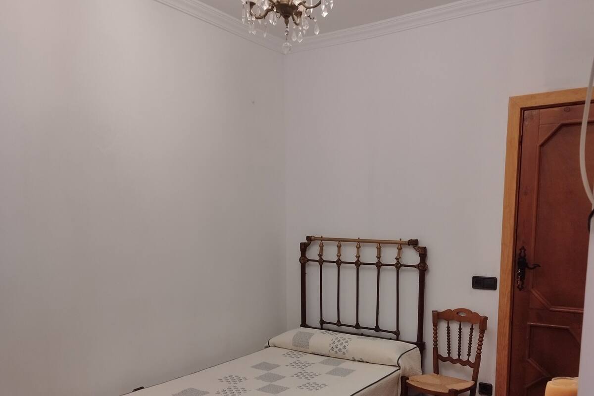 Une petite chambre à coucher, bien rangée, avec un lit simple, une chaise, une porte en bois et un lustre.