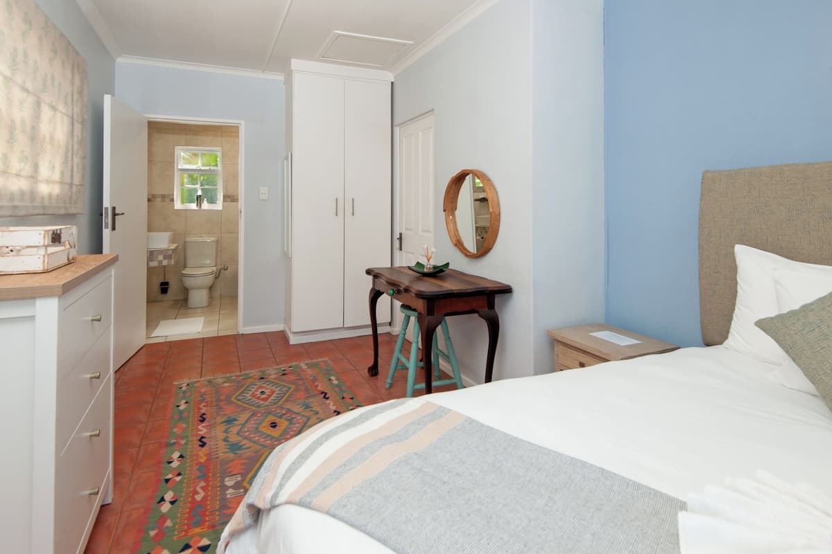 A bedroom with a bed, a nightstand, a wooden table with a mirror, a bathroom with a toilet, and a door leading to another room.