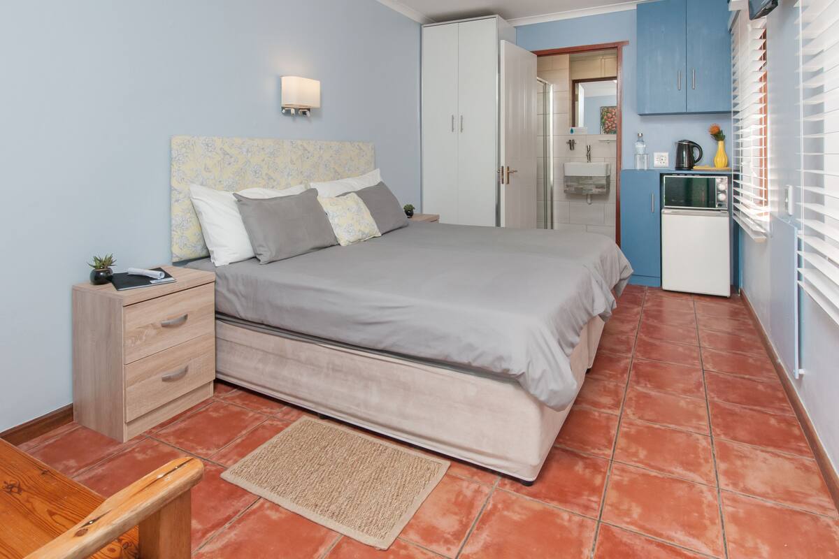 A bedroom with a bed, bedside table, and a small desk. There is a wardrobe, a kitchenette with a fridge, and a window with blinds.