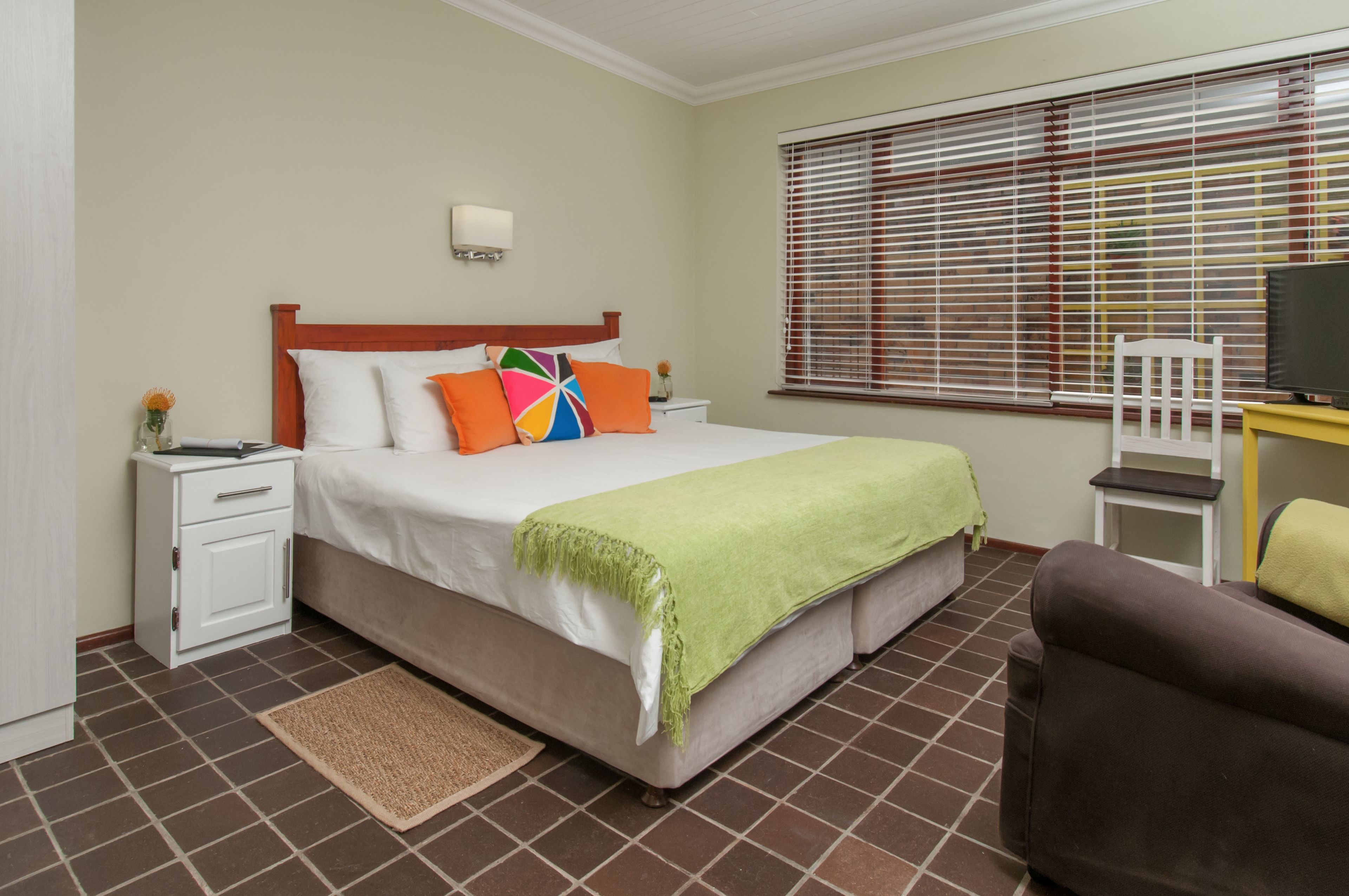 A hotel room with a bed, a nightstand, a chair, a TV, and a window with blinds.
