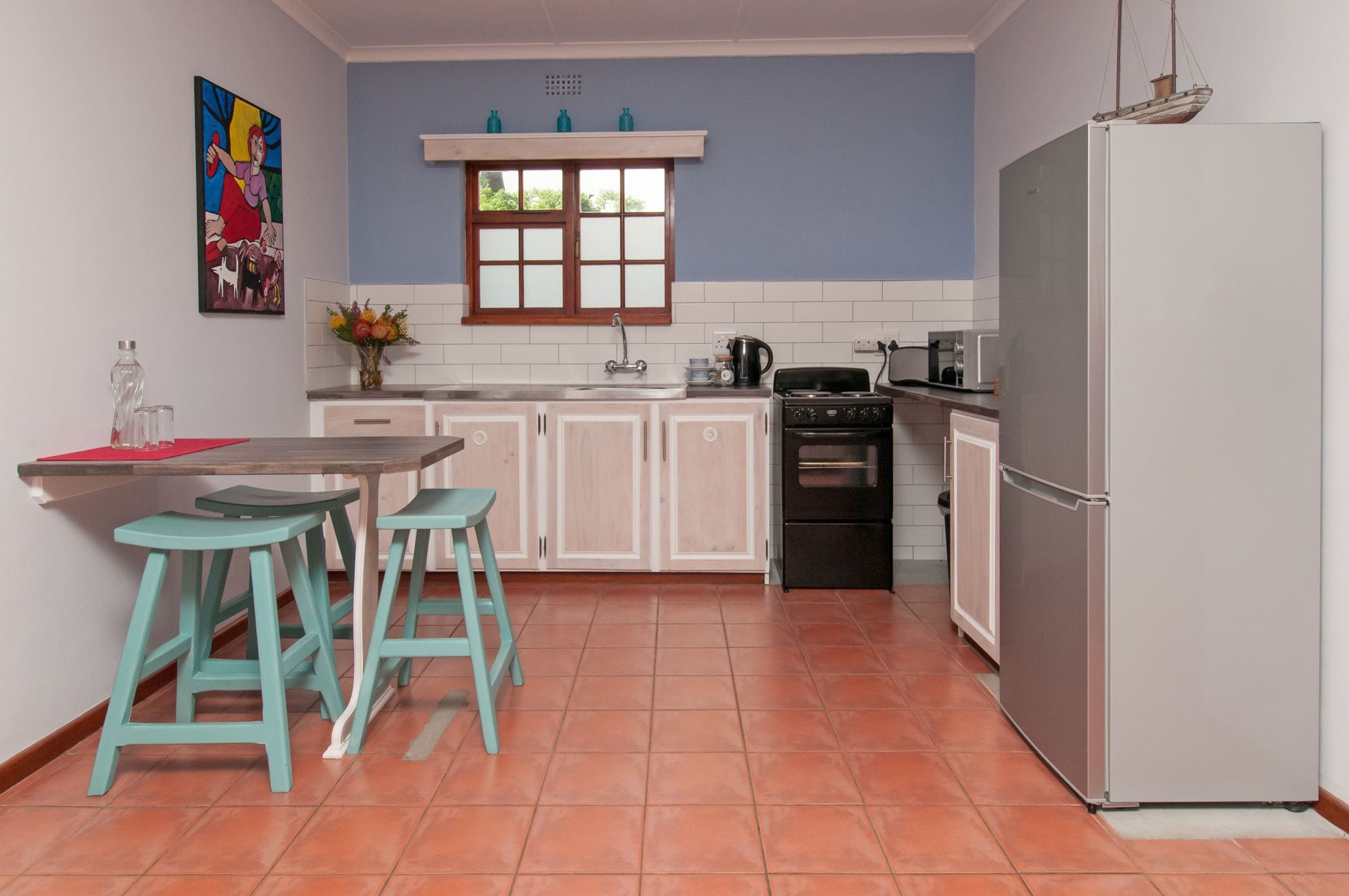 Een compacte keuken met witte kasten, een zwart fornuis en een witte koelkast. Er staan twee turquoise krukken bij het aanrecht.