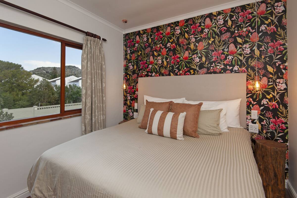 A bedroom with a large bed, floral wallpaper, a window with curtains, and a nightstand with a lamp.